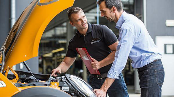 Zwei Männer inspizieren einen gelben MINI mit geöffnetem Motorraum in einer Werkstatt.