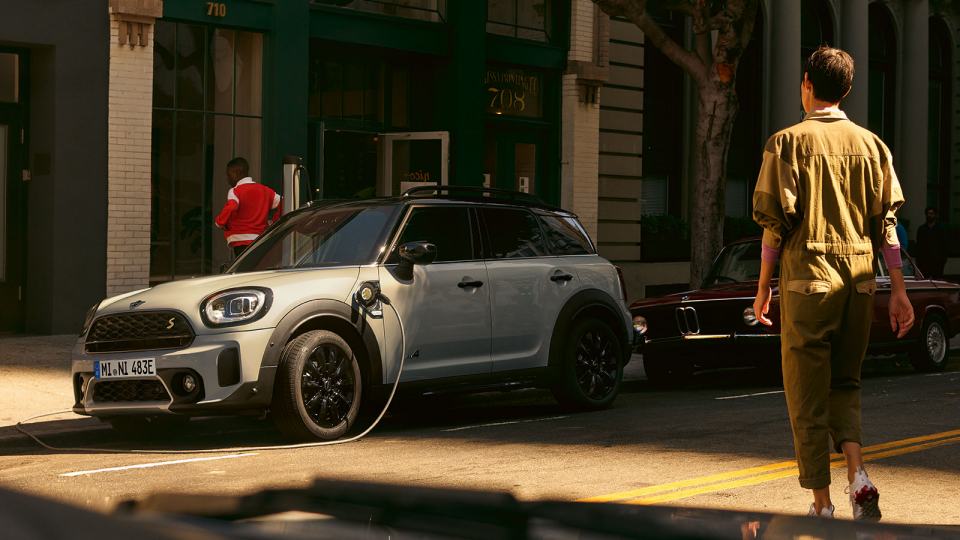 Graues Elektroauto an Ladestation, Person im Overall läuft vorbei, urbane Straßenszene.