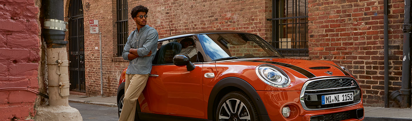 Mann lehnt an orangenem MINI vor Backsteinmauer; Stadtumgebung, entspannte Atmosphäre.