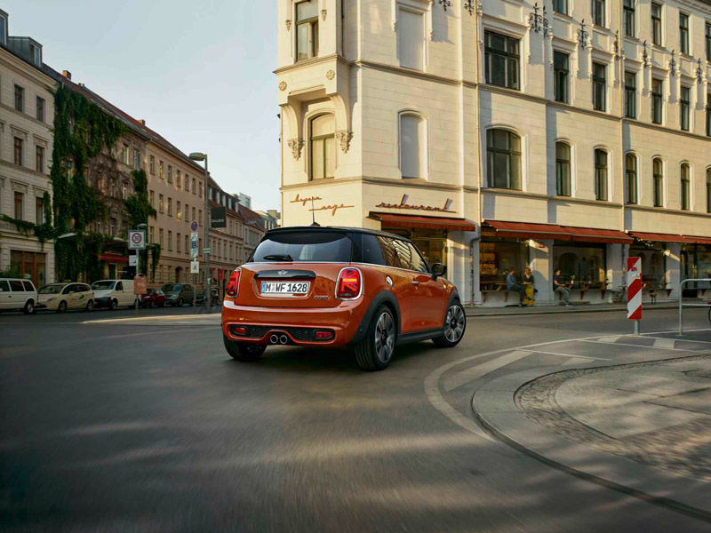 Roter MINI Cooper S fährt durch eine städtische Kurve vor einem großen weißen Gebäude.