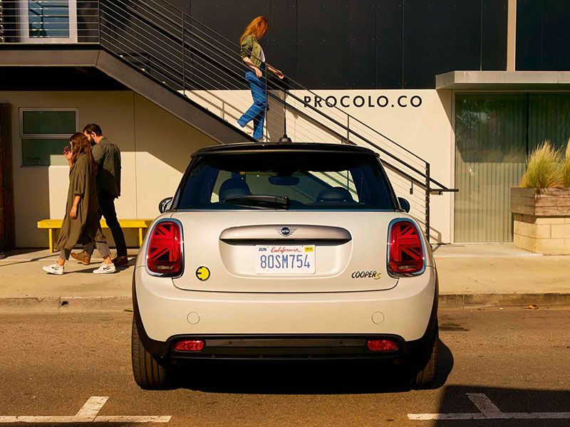 Weißer MINI Cooper SE, Heckansicht, geparkt vor modernem Gebäude mit Personen auf Gehweg.