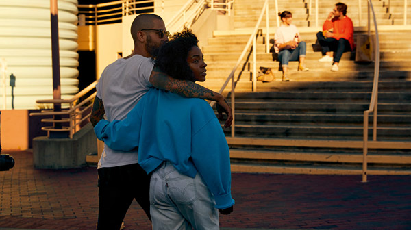 Ein Mann und eine Frau gehen eng umarmt auf einem Platz; im Hintergrund sitzen zwei Personen auf Stufen.
