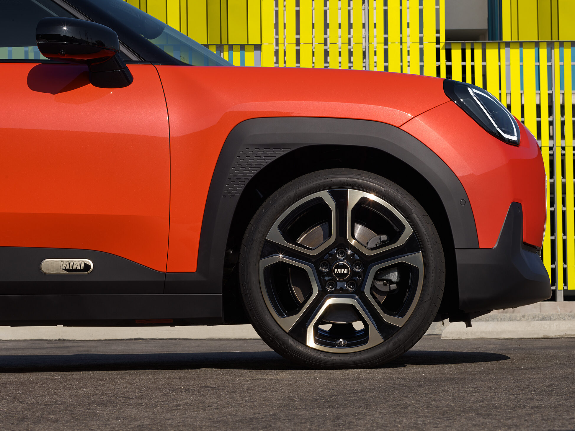 Roter MINI Cooper seitlich, mit schwarzem Spiegel und Felge, vor gelber Wand.