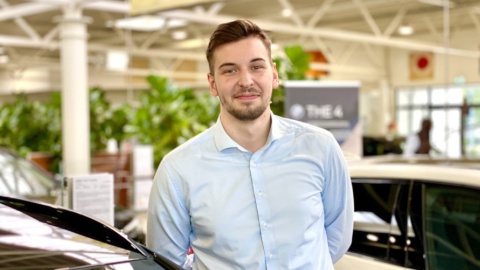 Ein Mann in einem hellblauen Hemd steht in einem Autohaus neben einem schwarzen Auto.