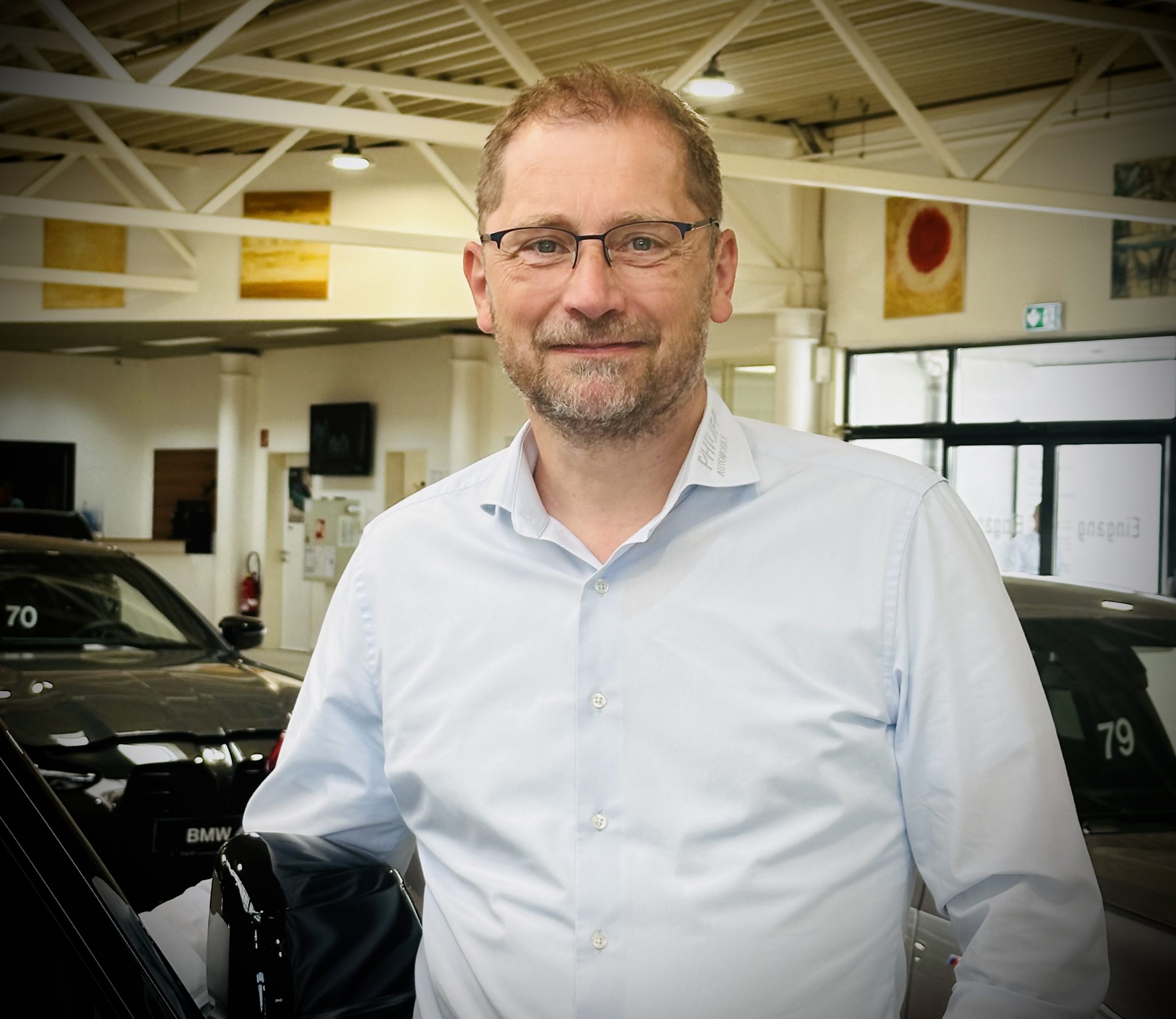 Mann mit Brille steht in einem Autohaus neben einem schwarzen Auto; im Hintergrund weitere Fahrzeuge.