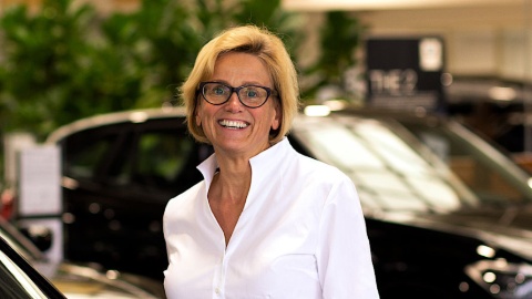 Eine lächelnde Frau in weißem Hemd vor unscharfen Autos im Innenraum.