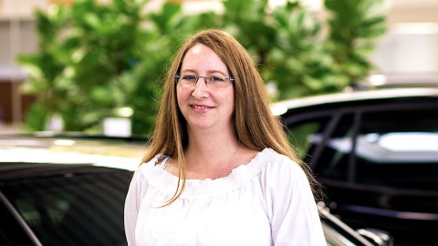 Eine Frau mit Brille steht lächelnd vor unscharfen Pflanzen und Autos im Hintergrund.