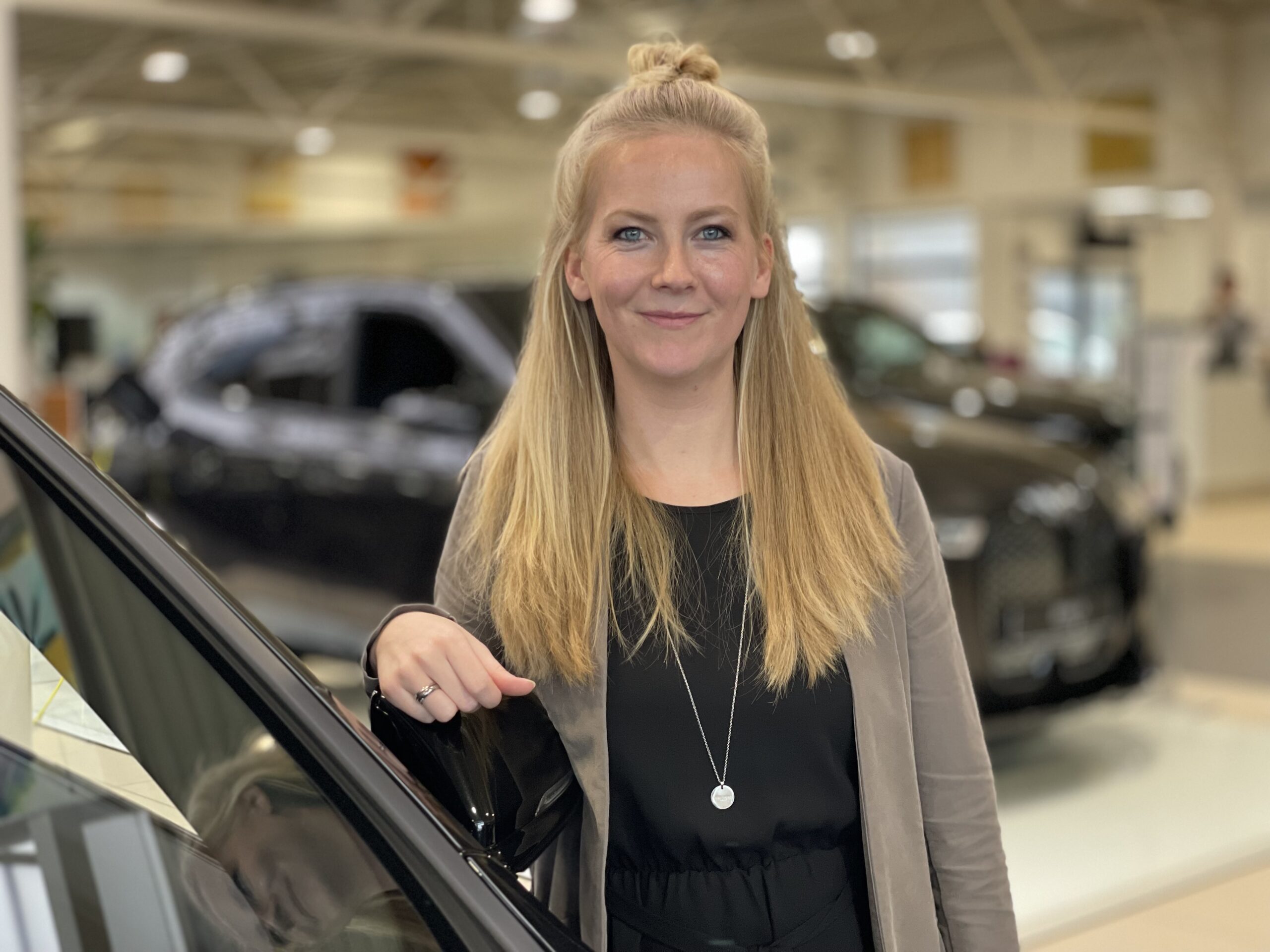Frau mit langen blonden Haaren steht in einem Autohaus neben einem Auto, lächelt in die Kamera.