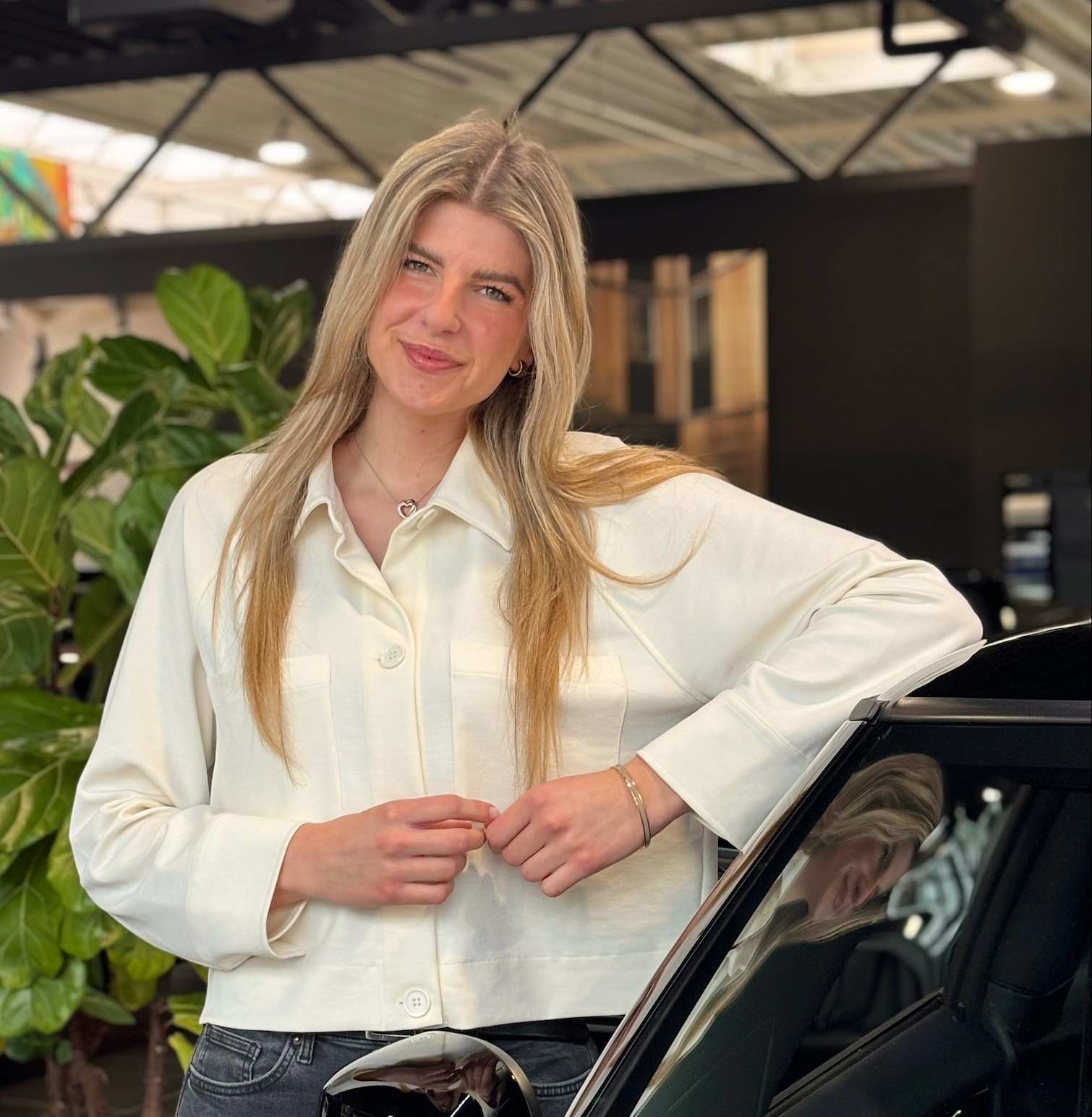 Eine lächelnde Person in einer weißen Bluse lehnt an einem schwarzen Auto im Ausstellungsraum.