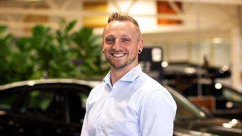 Ein Mann lächelt in einem Autohaus mit Pflanzen und Autos im Hintergrund.