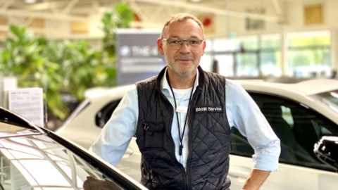 Eine Person in einem Autohaus steht neben einem Auto; im Hintergrund Pflanzen und Fahrzeuge.