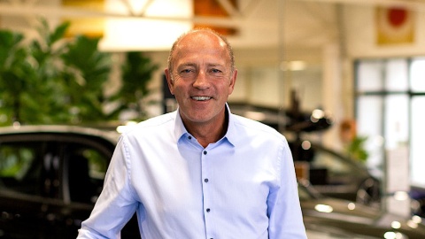 Mann in hellem Hemd steht in einem Autohaus mit unscharfen Autos im Hintergrund.