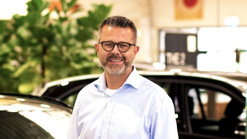 Ein lächelnder Mann mit Brille in einem Autohaus. Im Hintergrund Pflanzen und Autos.