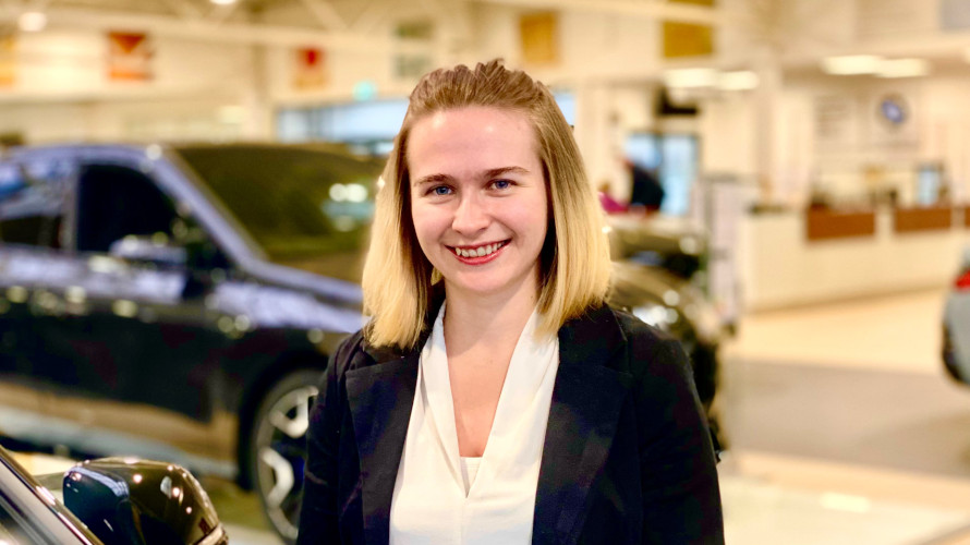 Eine lächelnde Frau in einem Autohaus mit verschwommenen Fahrzeugen im Hintergrund.