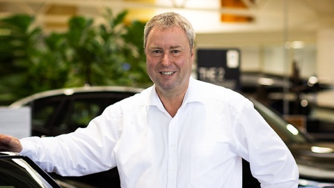 Ein Mann in weißem Hemd steht lächelnd vor einem Auto in einem Autohaus.