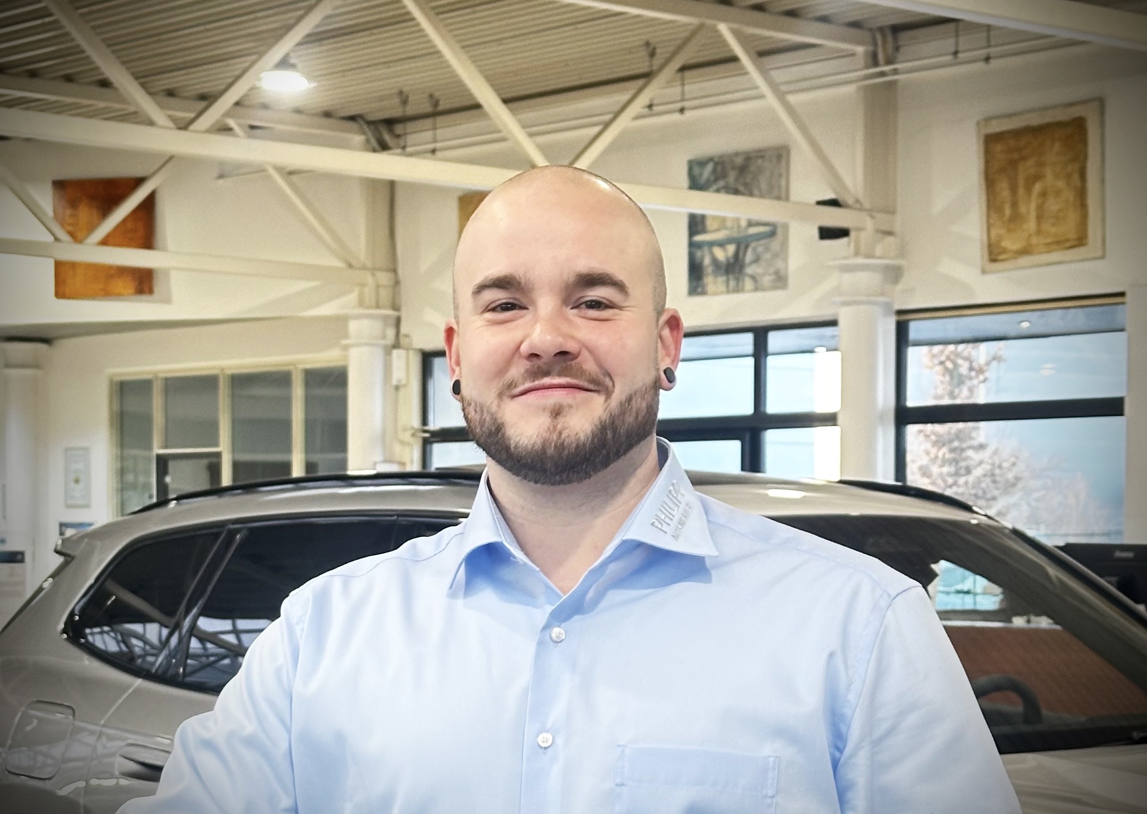 Ein Mann in hellblauem Hemd vor einem grauen Auto in einem Autohaus.